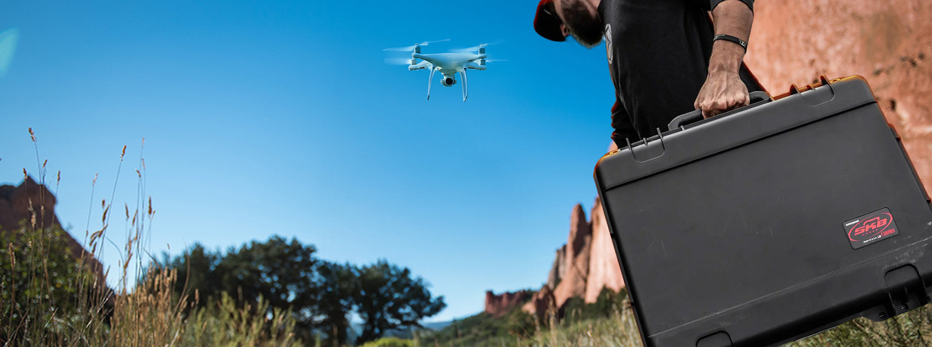 Person with a large black case and a drone flying in a scenic outdoor setting.