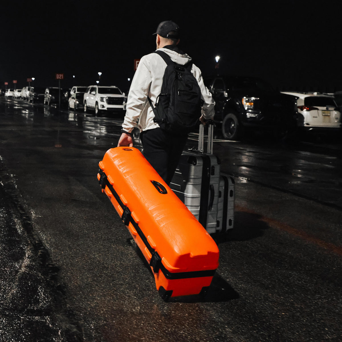 A man carrying a bright orange hard case and a silver rolling suitcase through a wet parking lot at night.