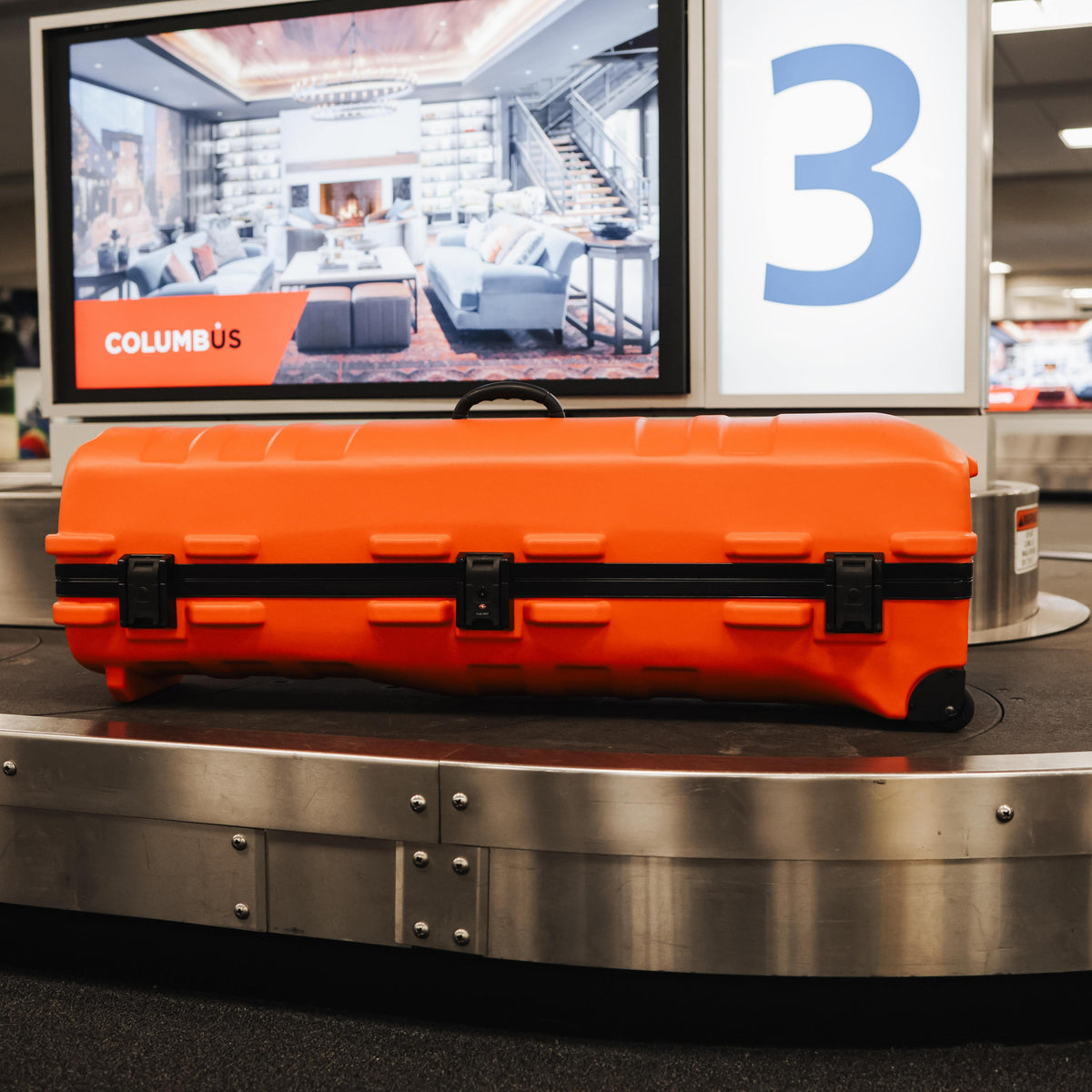 A bright orange hard-shell suitcase with black handles and trim sits on a metallic baggage carousel in an airport setting. A screen behind it shows a living room interior, and a large blue number 3 is visible on the signage.