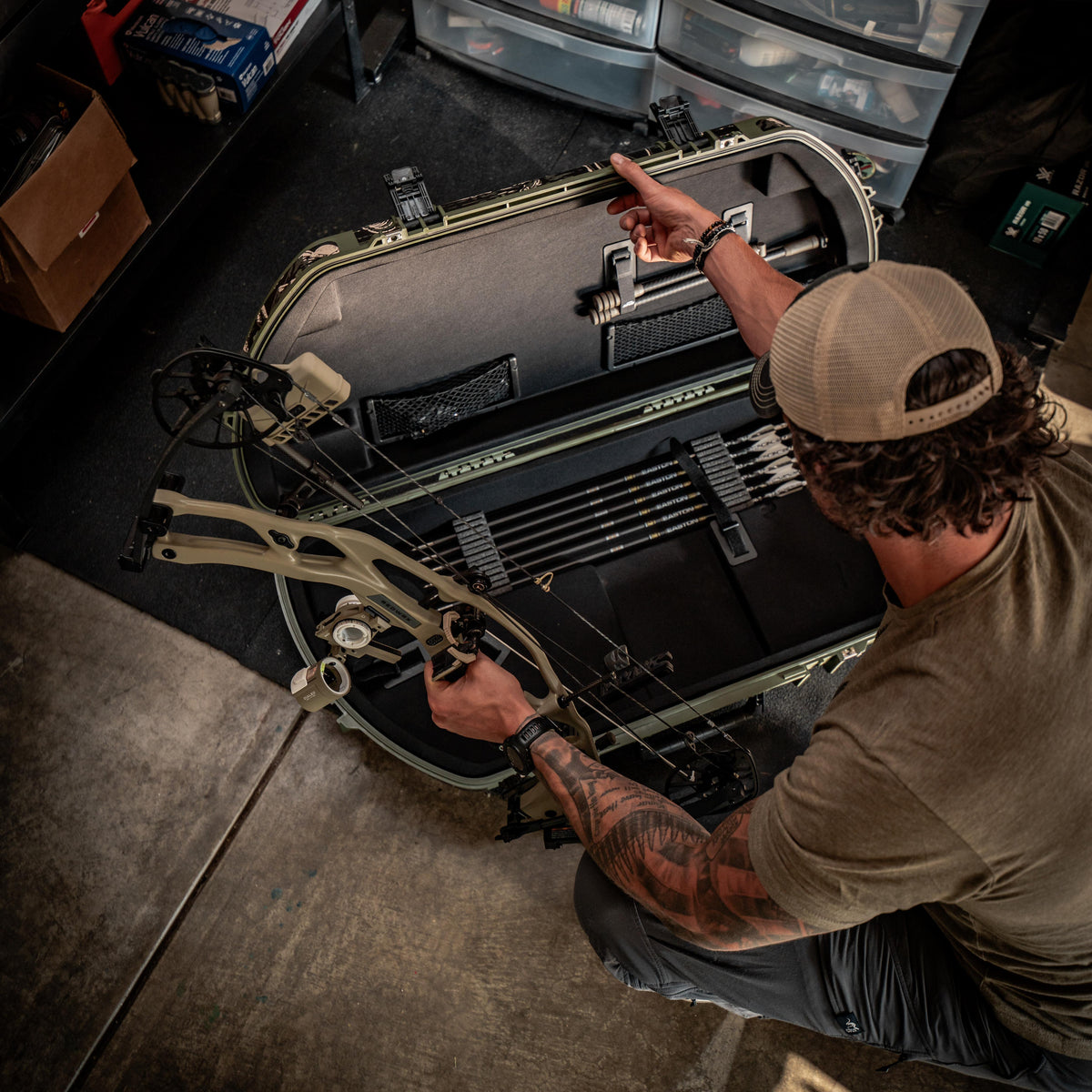 A man kneels beside an open hard case containing a compound bow and arrows, pointing inside the case in a storage room or workshop setting.