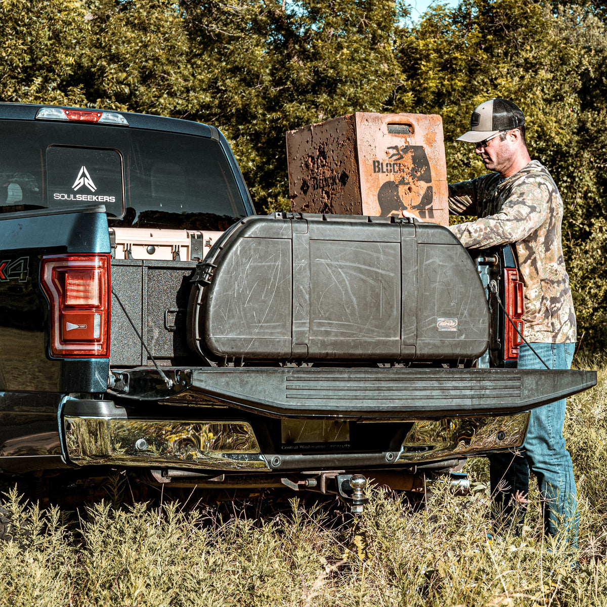 A man in camouflage clothing stands beside a black pickup truck in a grassy outdoor setting, holding a large black Soul Seekers hard case loaded in the truck bed next to a rusted wooden crate.