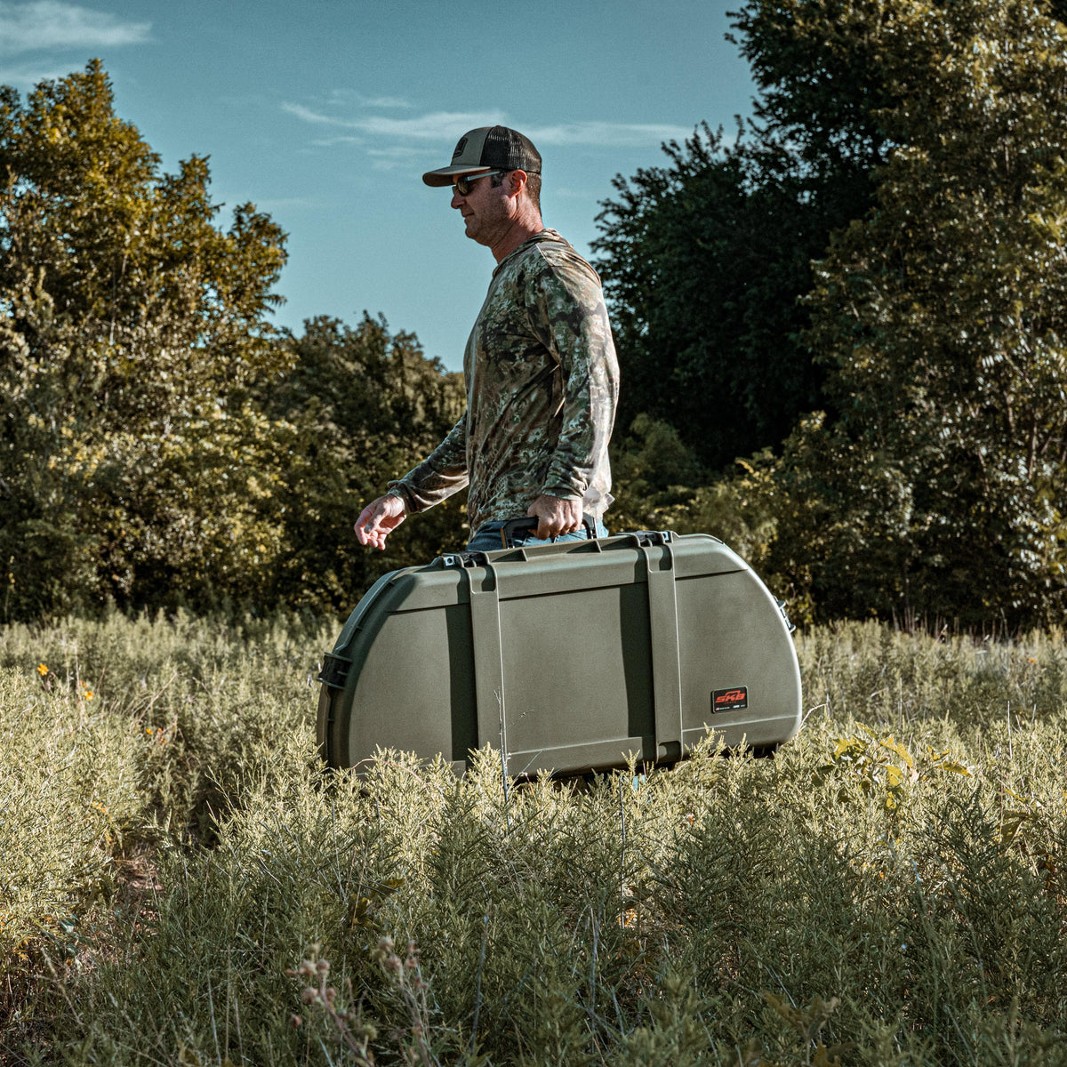 A man in a camouflage long-sleeve shirt and cap carries a large olive green hard case outdoors in a grassy field with trees in the background.