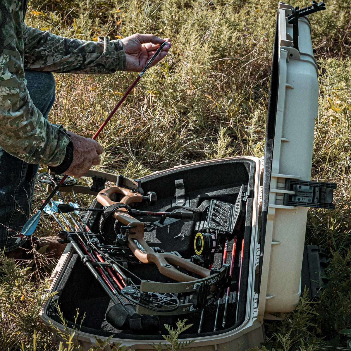 An open hard case filled with archery equipment, including bows and arrows, set in outdoor grassy terrain. A person in camouflage clothing is partially visible on the left, handling an arrow.
