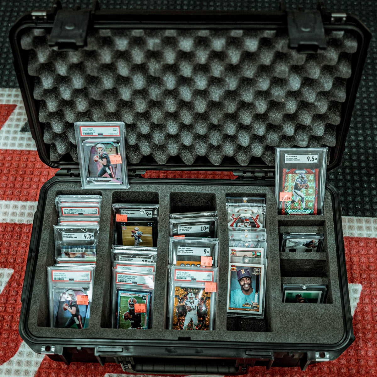 An open black hard case with custom foam inserts, filled with multiple graded and encapsulated sports trading cards. Several cards show visible PSA grading labels, and small orange price tags are placed on some cards.