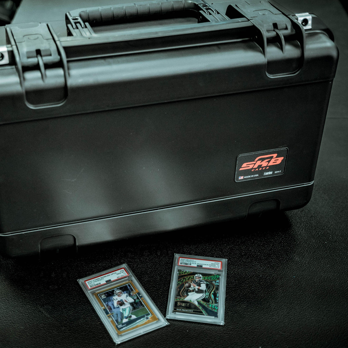 A black hard protective case with a handle and a visible SMB logo patch, photographed on a dark surface. Two graded trading cards are placed in front of the case.