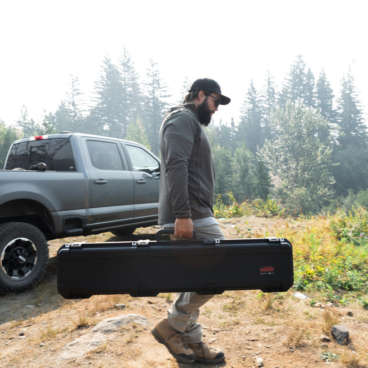 A man carrying a large black hard case outdoors, walking beside a gray pickup truck in a wooded, grassy area. The case has a small red and white logo near the lower right end.