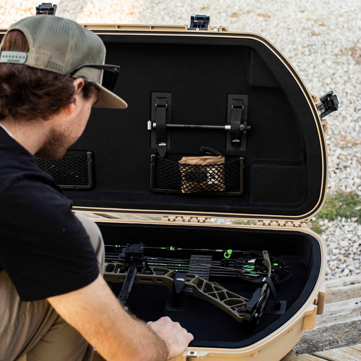 A man in a black t-shirt and cap looks into an open tan hard case containing a compound bow and accessories, set outdoors on a wooden surface with gravel in the background.