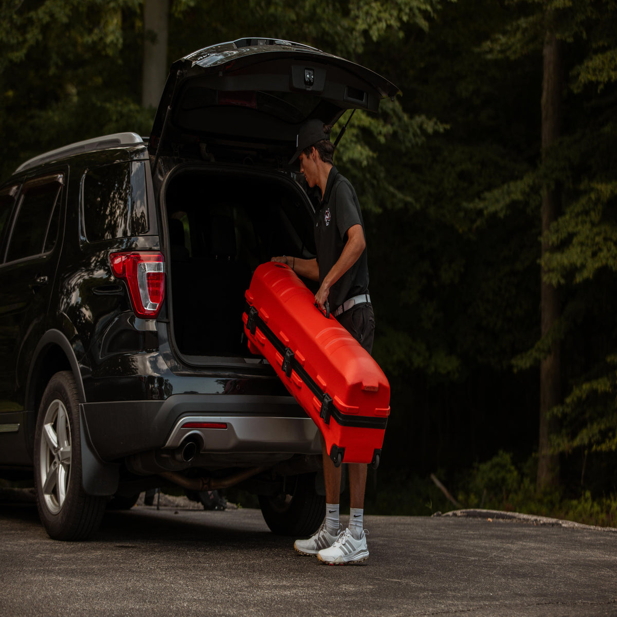 A man in a black shirt and cap carries a bright red hard case into the open trunk of a black SUV, with a forest background.