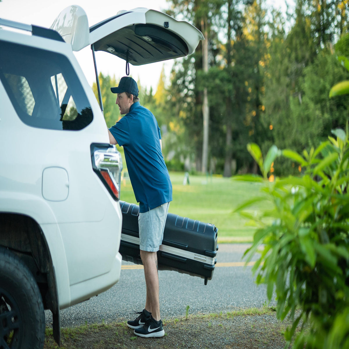 A man in a blue shirt and gray shorts loads a black travel case into the trunk of a white SUV outdoors on a paved road with trees in the background.