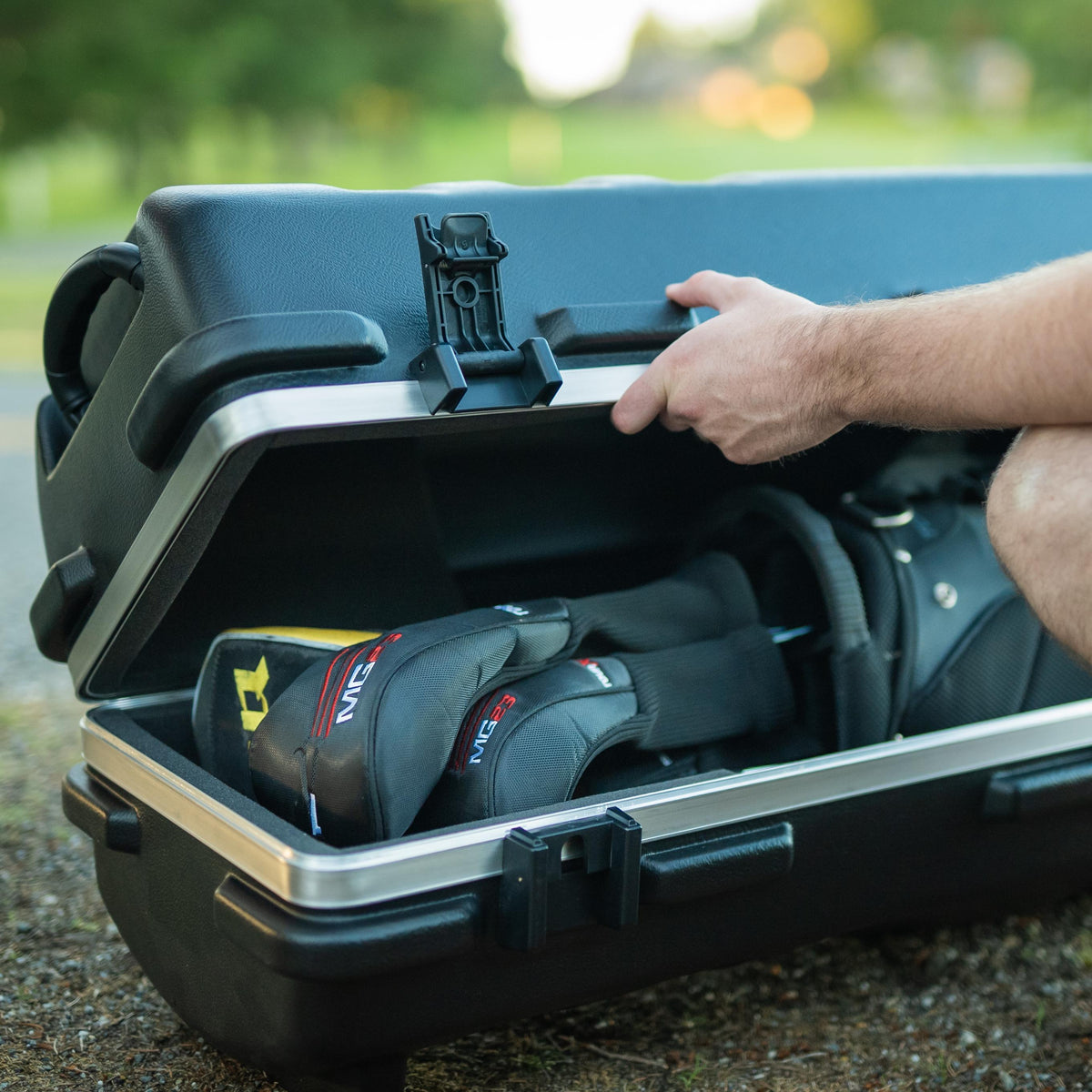 A black hard-shell storage case is open on a gravel surface, with a person's hand and leg visible as they place or retrieve gear inside. The case has silver metal trim and latches, and the interior contains black gear bags with red and yellow branding.