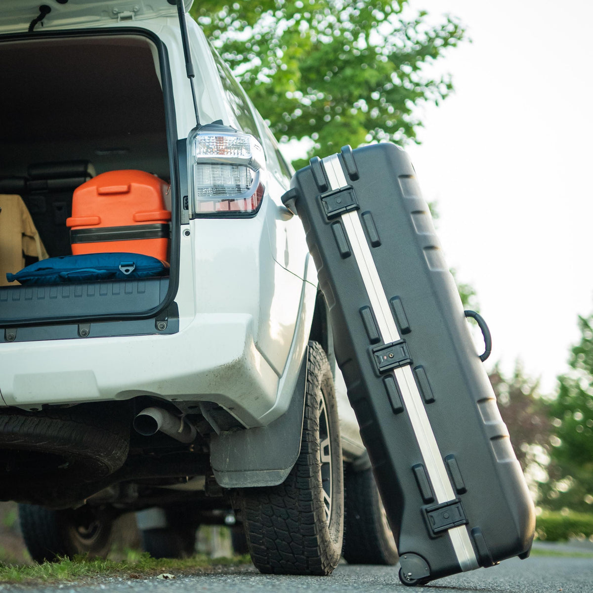 A black hard-shell travel case with silver accents and a handle, leaning against the open trunk of a white vehicle outdoors.