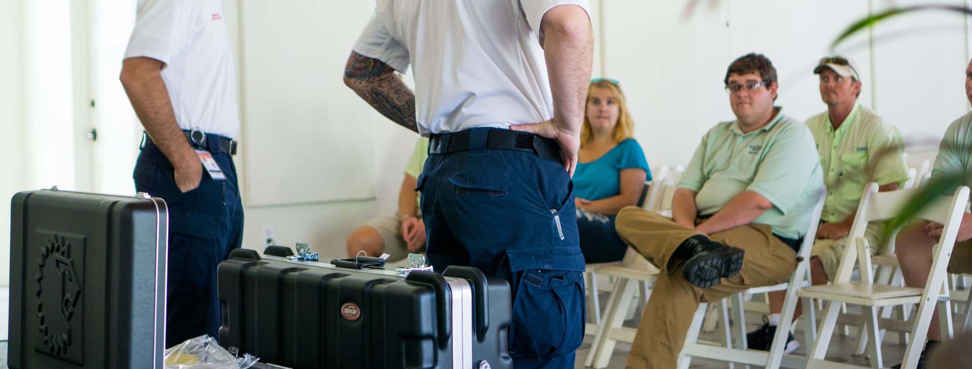 Person in a white shirt and blue pants standing next to audio equipment with people sitting in the background.