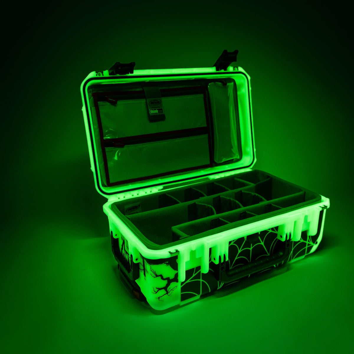 A hard-sided storage case with an open lid, shown glowing green in the dark. The case has molded organizer compartments inside and a black spiderweb graphic on the front panel.