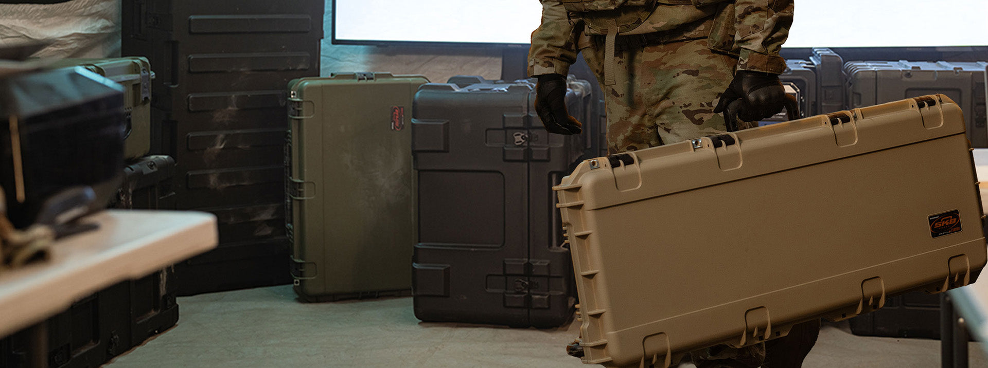 Person in camouflage holding a large beige hard-shell case in a room with other storage cases.
