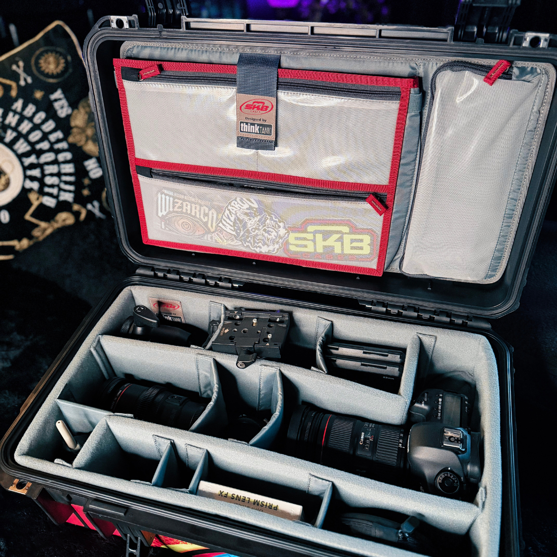 An open black hard camera case with gray foam interior compartments, shown holding lenses and camera gear. The inside lid has red-trimmed pockets and a visible DigiRack SKB tag.
