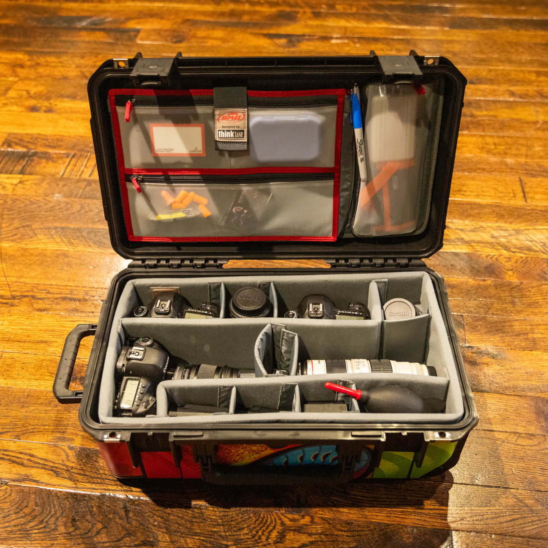 An open black hard-shell camera gear case on a wooden floor, filled with camera accessories and organized in foam compartments.
