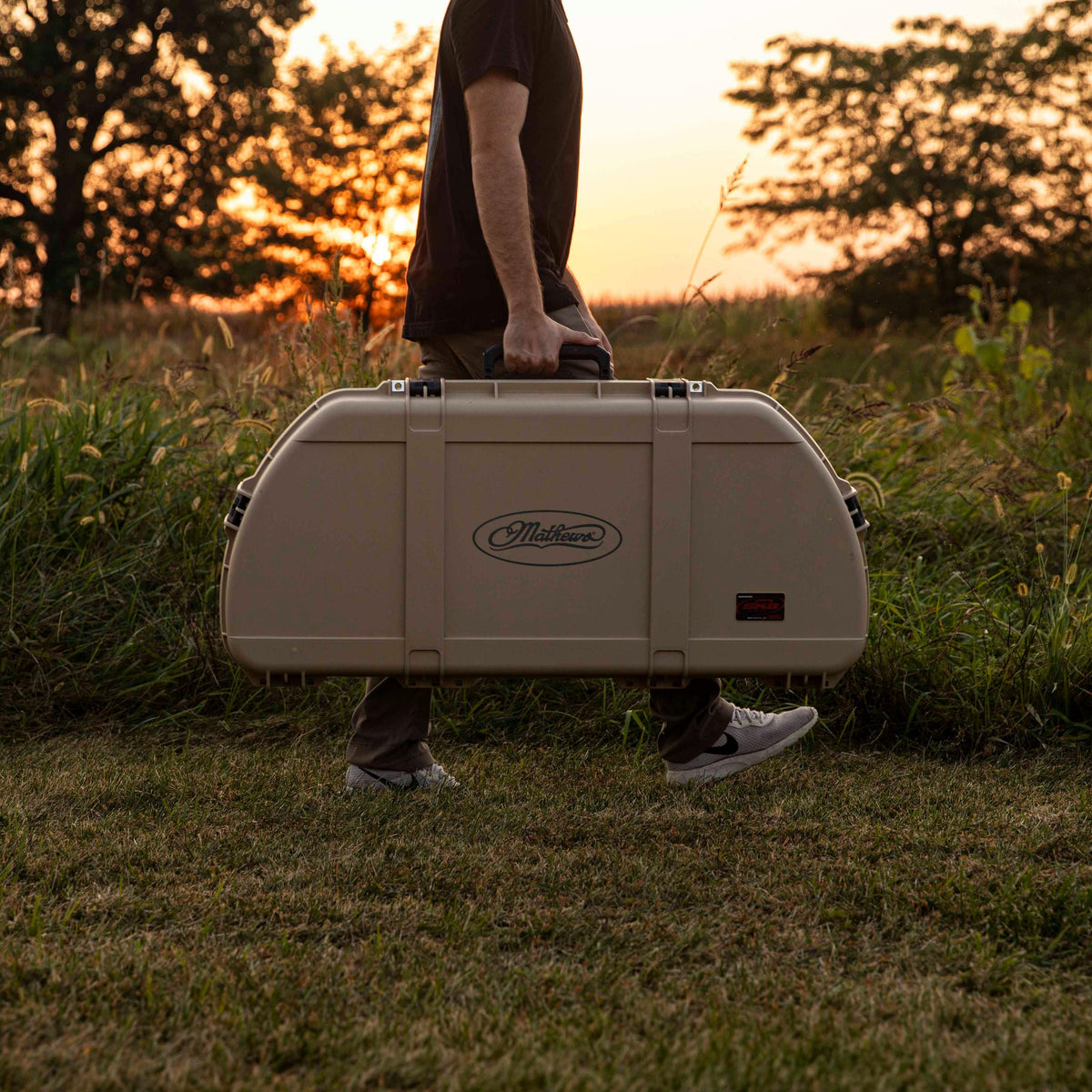 A person carrying a large beige hard case outdoors in a grassy field at sunset. The case has a visible 'Maldonado' logo on the front and is shown in use with trees and warm golden light in the background.